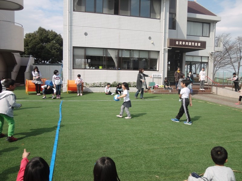 晴天の下で佐賀市川副児童館の芝生広場に集まった大勢の子供たちが、青いラインで区切られたコート内でボール遊びを楽しみ、背後の白いタイル貼りの施設で見守る大人たちと共に活動しているドッジボール大会の様子の写真