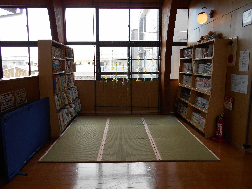 大きな窓から光が差し込む一角に畳スペースがあり、左右の本棚に絵本や児童書が整然と並ぶ落ち着いた図書コーナーの写真