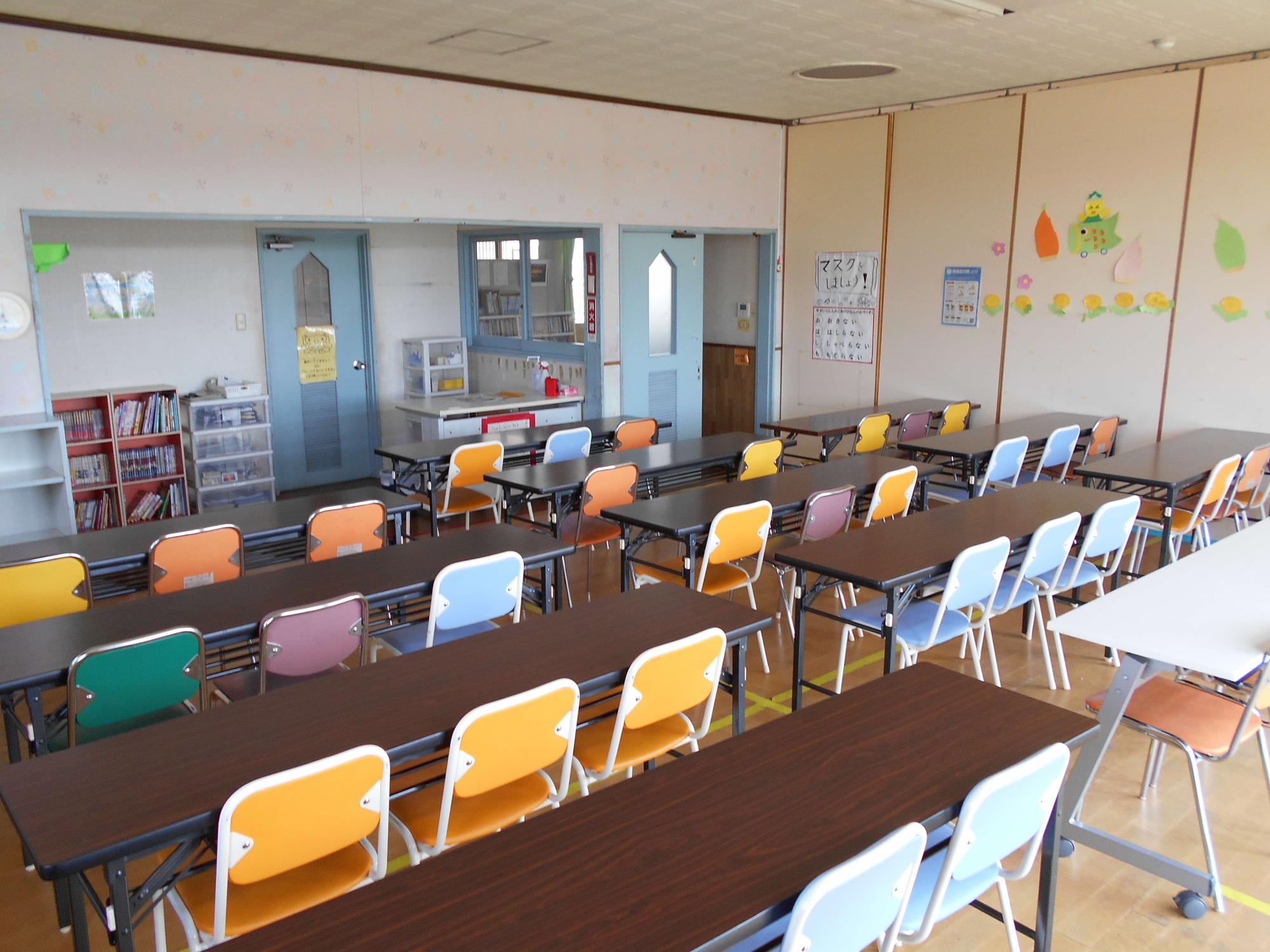 色とりどりの椅子と長机が整然と並ぶ広い児童クラブ室で、壁面装飾や本棚、青い扉が温かで落ち着いた学習環境を感じさせる写真