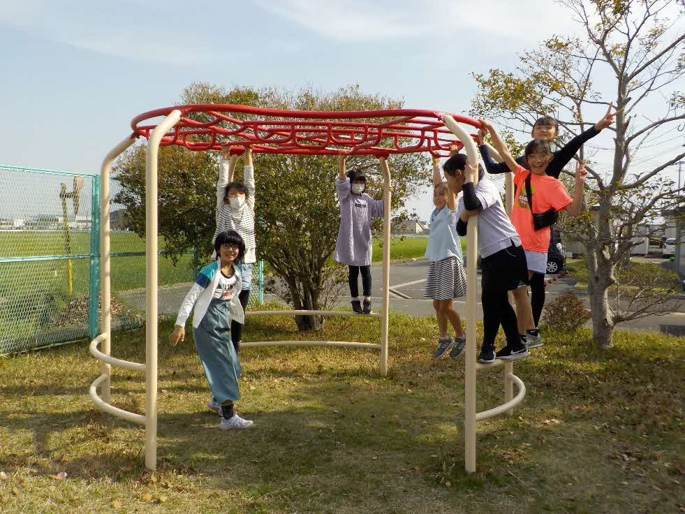 屋外の公園にて、赤い円形の雲梯が備わった白いフレームの宇宙船型ラダーに複数の子供たちがぶら下がったり登ったりして遊び、カメラに向かって笑顔でポーズをとっている写真