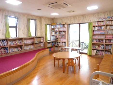 大きな窓から光が差し込む室内に本棚が壁一面に並び、丸い机と椅子が配置された明るい図書コーナーの写真