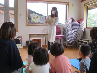 室内で大人が絵本を掲げて読み聞かせを行い子どもたちが床に座って熱心に見入る様子の写真