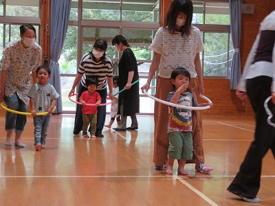 広い窓のある明るい体育館で子どもたちが大人と一緒にフラフープを使って歩いたり遊んだりする様子を写した写真