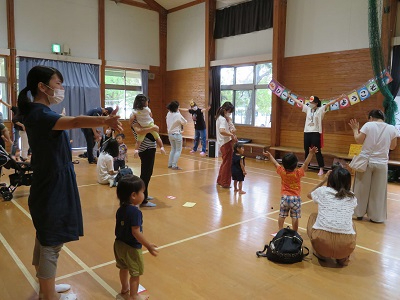 明るい体育館で子どもと大人が輪になって体を動かし講師の動きをまねて楽しむ様子の写真