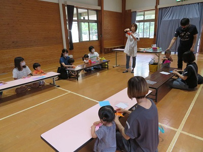 明るい室内で子どもと大人が低い机に向かって折り紙などの制作を行い指導者が手本を示している写真