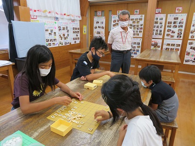 木の机を囲んで子どもたちが将棋の駒を並べて対局し、大人が静かに見守る室内の活動風景の写真