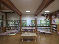 木の梁がある室内で壁一面の本棚と掲示物に囲まれた読書コーナーに2台の木製のテーブルと、左右にL字型の低い机が並ぶ写真