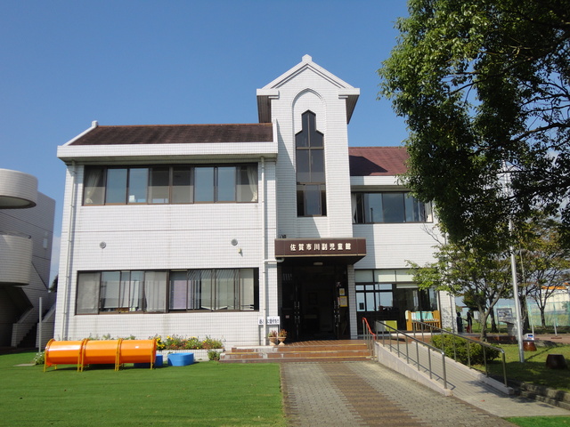 晴天の下、白いタイル壁に茶色の屋根を冠した佐賀市川副児童館の外観が写っており、芝生の庭にはオレンジ色のトンネル状の遊具が設置され、館内へ続くスロープと階段が入り口に繋がっている施設の建物外観の写真