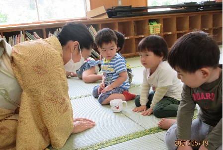 着物を着てマスクをした女性が、座っている幼い子供たちの前で丁寧に両手を突いてお辞儀をしており、子供たちがその様子を見ながら同じようにお辞儀をしている茶道体験の写真