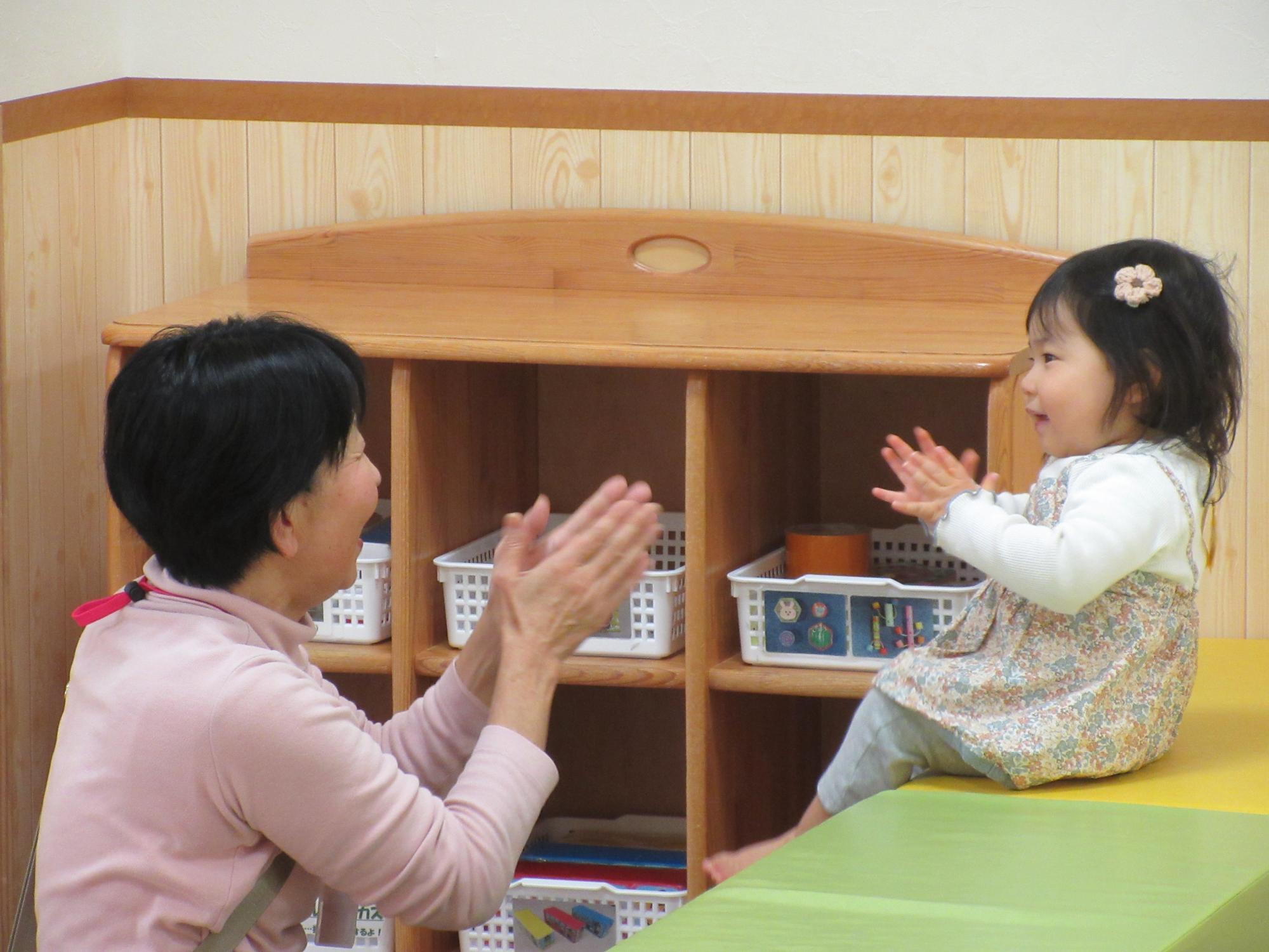 明るい保育室で木製棚の前に座った大人と幼い女の子が向かい合い楽しげに手を叩き合いながら穏やかに過ごす様子の写真