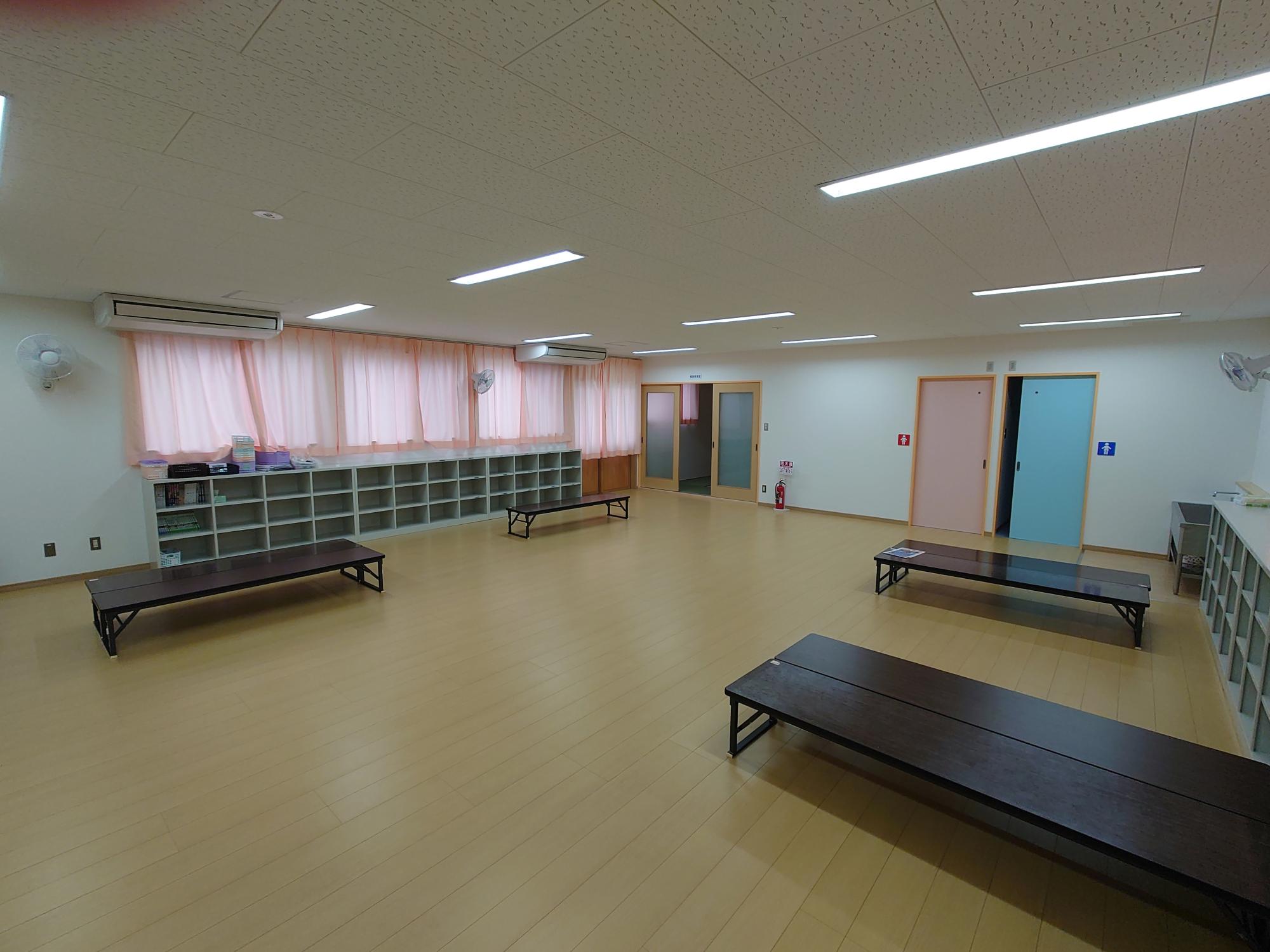 ベージュのフローリング床に低い長机が配置され、壁際に収納棚とピンクのカーテンがある児童クラブの室内写真