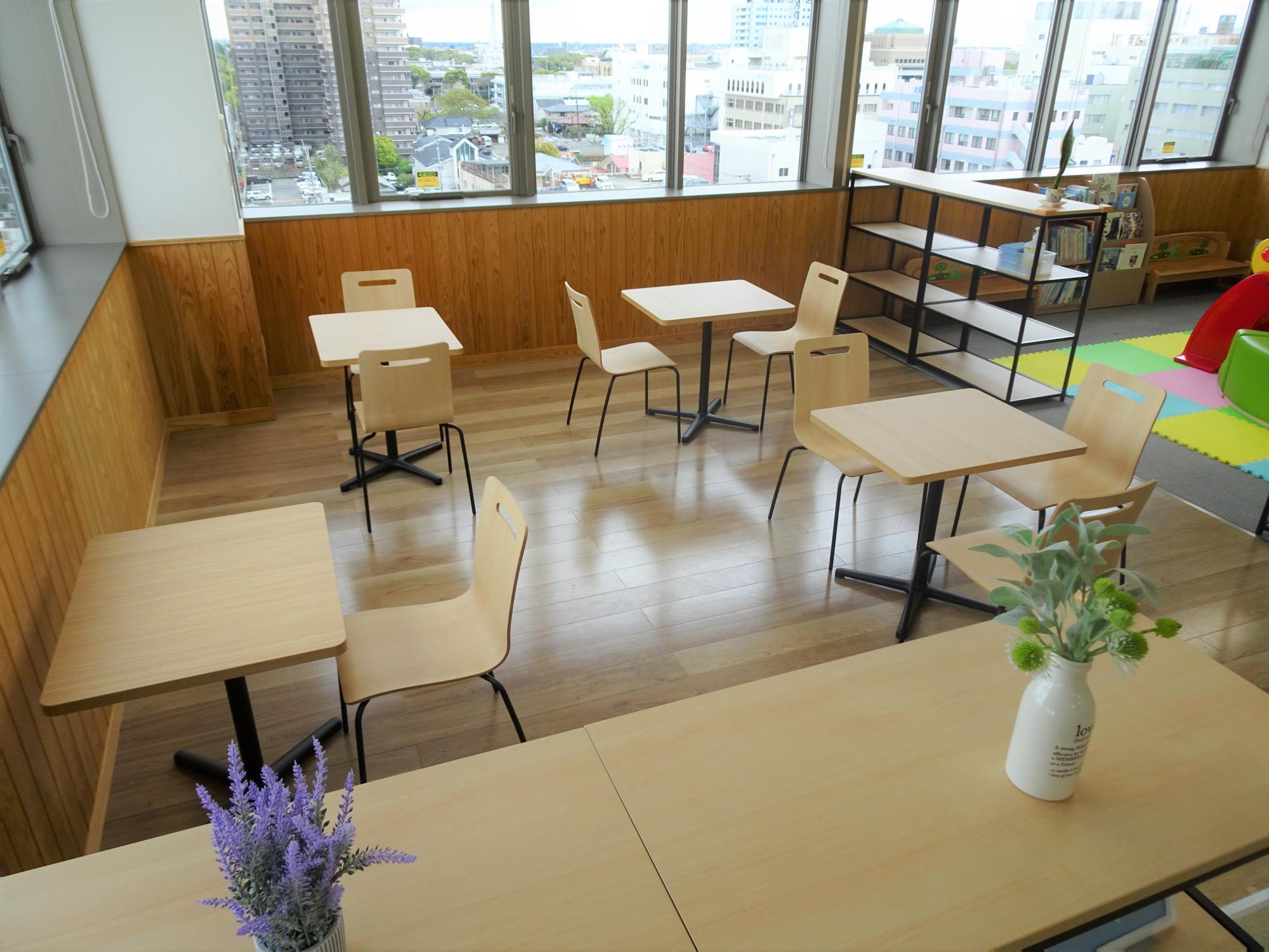 大きな窓から街並みが見える明るい室内で、木製テーブルと椅子がゆったり配置され、棚や遊具も備える空間の写真