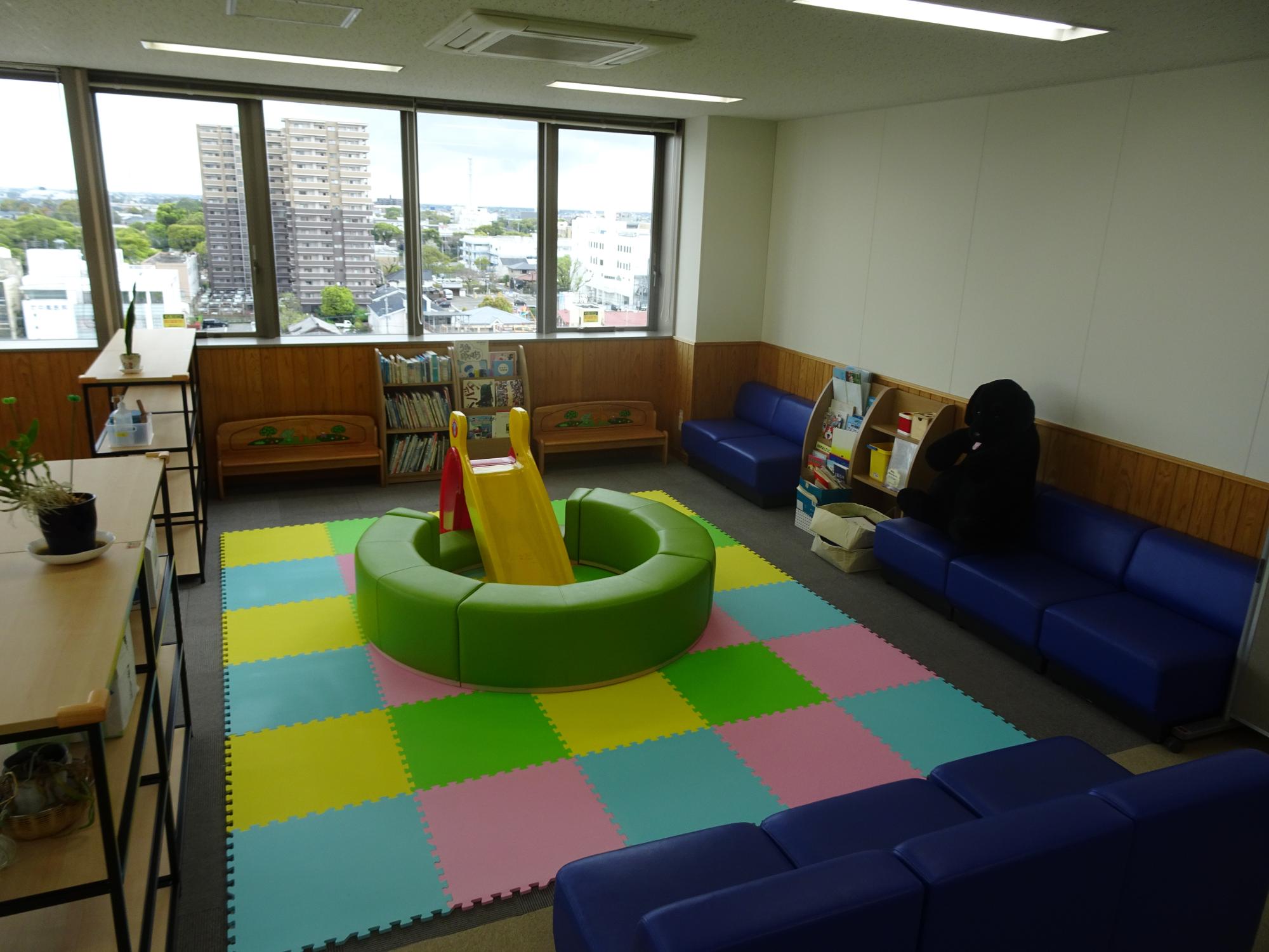 大きな窓のある明るい室内で色とりどりのマット中央に滑り台が置かれ周囲を青いソファがゆったり囲むキッズコーナーの写真