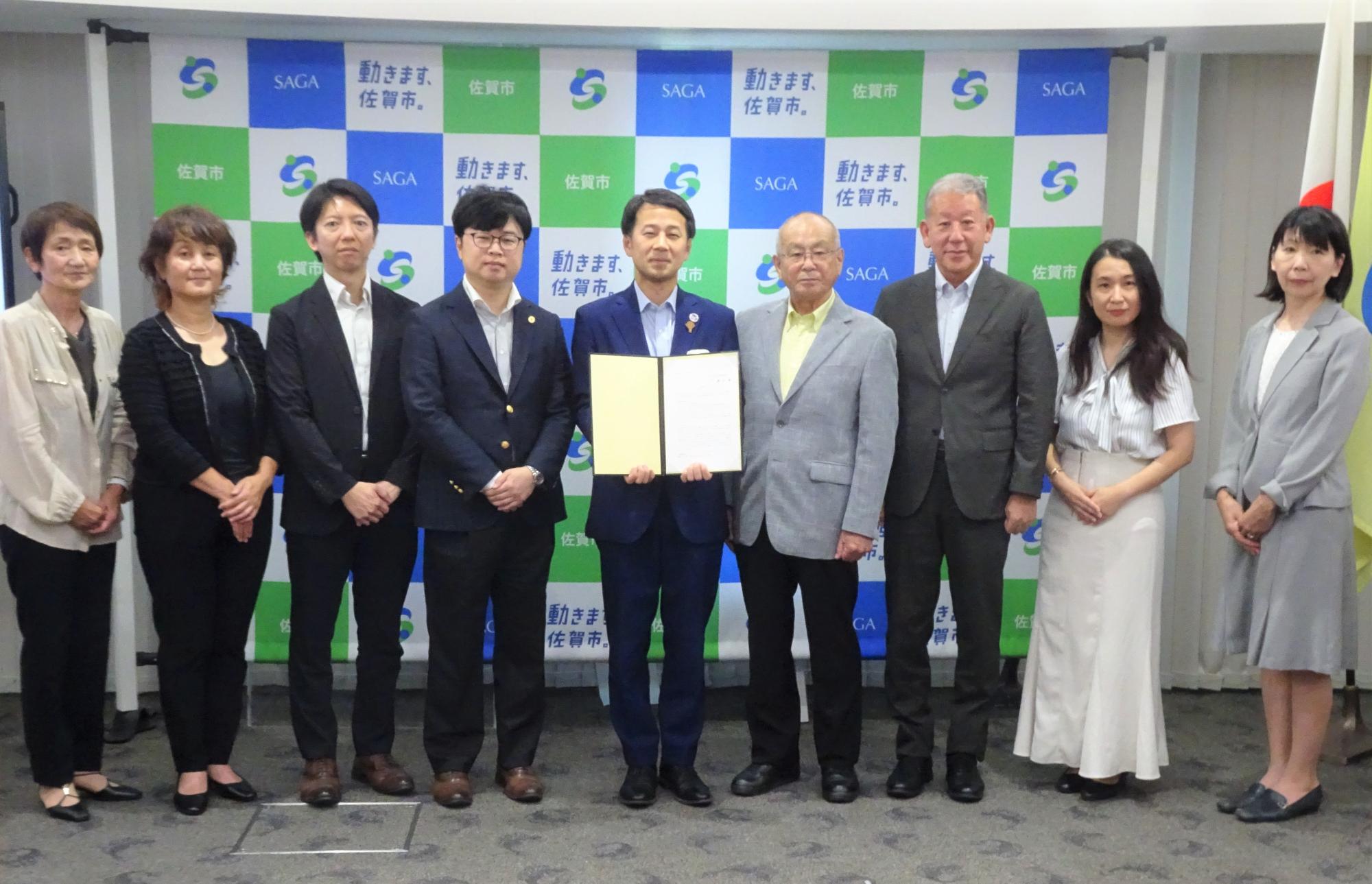 佐賀市のロゴが並ぶ背景の前で、答申書を持った市長を中心に複数の参加者が横一列に並び、記念撮影を行っている写真