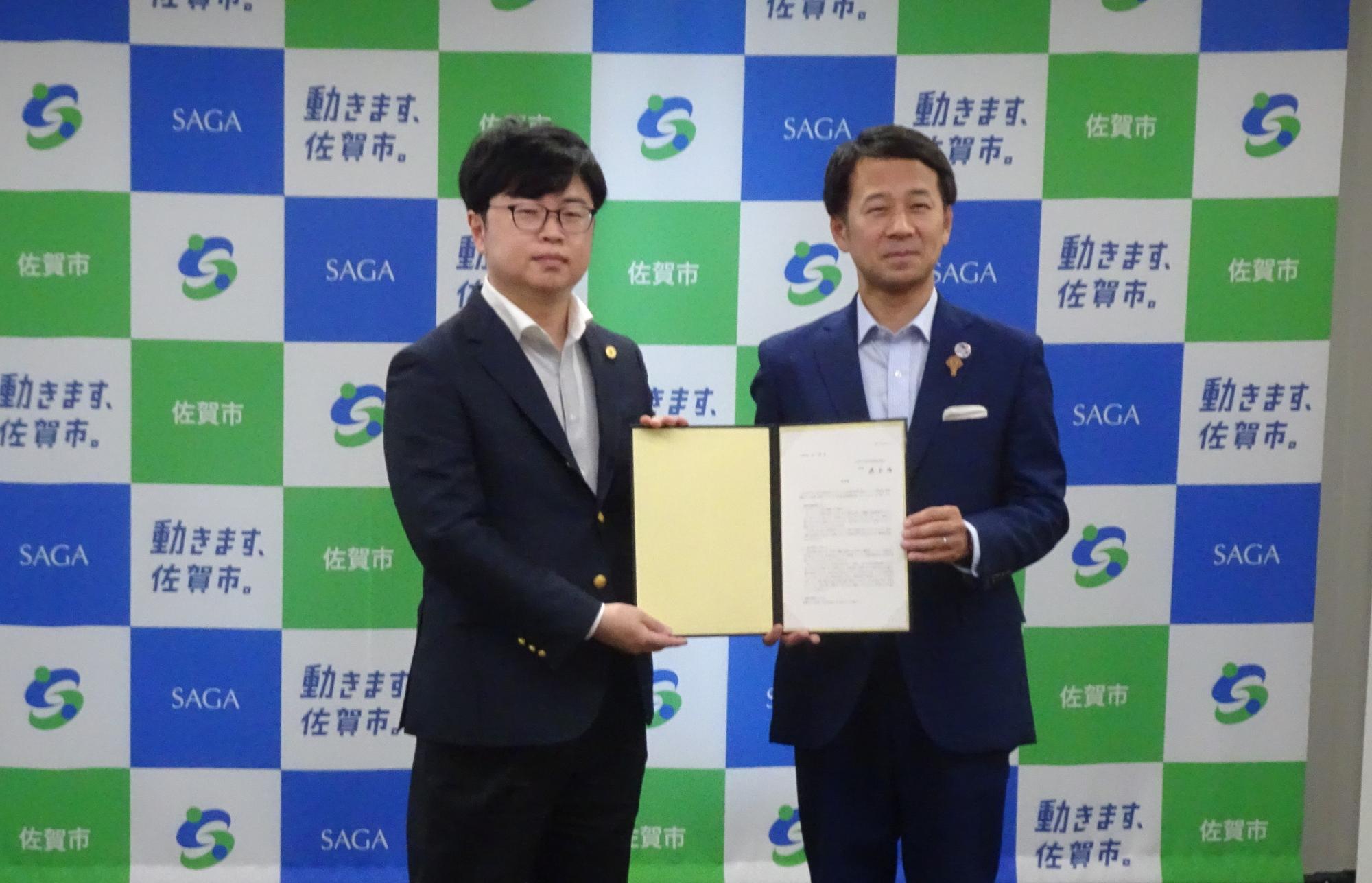 佐賀市ロゴが規則的に並ぶ明るい背景の前で男性と市長が答申書を共同でしっかりと掲げ記念撮影に臨む場面の写真