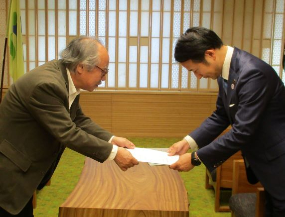 和風の格子窓がある落ち着いた室内で、ジャケット姿の五十嵐勉委員長とスーツ姿の市長が、互いに深く腰を折り曲げて丁寧に両手で提言書を受け渡している様子の写真