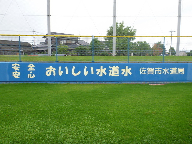 青々とした芝生が広がる球場の外野フェンスに、佐賀市水道局による「安全 安心 おいしい水道水」という白い文字の広告が青い背景に大きく掲げられた写真