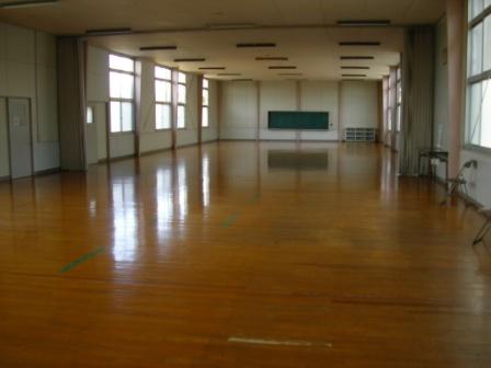 窓から自然光が入り込む広々とした多目的ホールで、磨き上げられた木製フローリングが光を反射し、正面奥の壁には大きな黒板が設置された、スポーツルームの写真