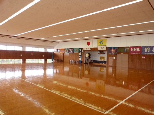 光沢のある茶色の床が広がる武道場の内部で、正面の壁面には日本の国旗や太鼓、多くの横断幕が掲げられており、左側の窓から差し込む光が床に反射し、天井の長い照明とともに神聖な空気が漂う武道場を捉えた写真