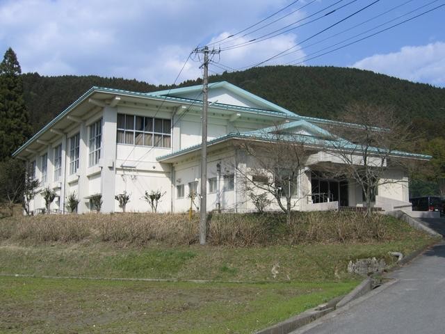 緑色の山並みを背に建つ白壁と薄い青色の屋根が特徴的な三瀬勤労福祉センターを、電柱や枯れ草の茂る斜面越しに斜め前方から捉えた写真