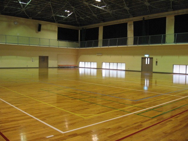 天井の高い鉄骨トラス構造の屋内体育館において、光沢のある木製フローリングの床面にバスケットボールやバドミントンなどの競技用ラインが引かれ、壁際には観覧用バルコニーが設置された無人の広々とした空間を写した写真