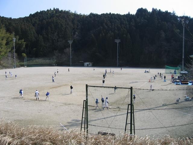 山あいに位置する夜間照明付きの広い土のグラウンドで、ユニフォーム姿の人々が集まって野球の試合を行っており、手前には防球ネットが設置され、背景には深い緑の山々が広がる写真