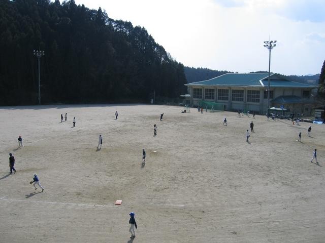 山あいの静かな環境にある照明設備付きの広い土のグラウンドで、大勢の選手たちがユニフォーム姿で野球の練習に励んでおり、背景には深い森と体育館のような建物が見える、活気あるスポーツの風景を収めた写真