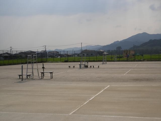 遠くに山々を望むのどかな風景の中、審判台やベンチが設置された複数面のクレーコートが広がり、周囲をフェンスで囲まれた開放感のある屋外テニスコートを、コートのラインが手前に見える角度から捉えた写真