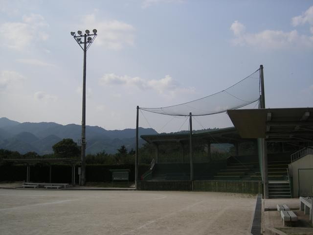 空には薄い雲が広がる山脈を背景にした屋外の野球場において、バックネット裏の屋根付き観客席と高い照明塔が設置されたグラウンドの様子を捉えている、自由広場の写真