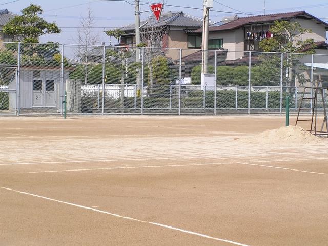 住宅街に隣接した土のテニスコートを捉えており、手前には白いラインが引かれたコートが広がり、奥にはメッシュフェンスがある諸富公園体育施設の景写