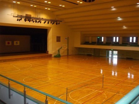 照明が灯る広々とした体育館の内部において、光沢のある木製フロアにはバスケットボールやバドミントンのコートラインが引かれ、奥には舞台やバスケットゴール、2階の観覧席までを見渡せるように高い位置から撮影した写真