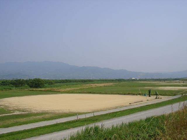青い空と遠くの山々を背景に、手前には未舗装の道路が走り、その奥に広がる緑豊かな河川敷に砂地で整備されたグラウンドや広場が点在している、のどかな風景を捉えた引きの構図で撮影された嘉瀬川河川敷北グラウンドの写真