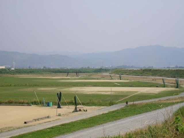 薄く霞んだ青空の下、遠くに連なる山々を背景として、手前に未舗装の道路、その奥に緑の草地と砂地のグラウンドが広がる河川敷の場所で、防球ネットなどの設備が点在するのどかな風景を捉えた嘉瀬川河川敷北グラウンド写真
