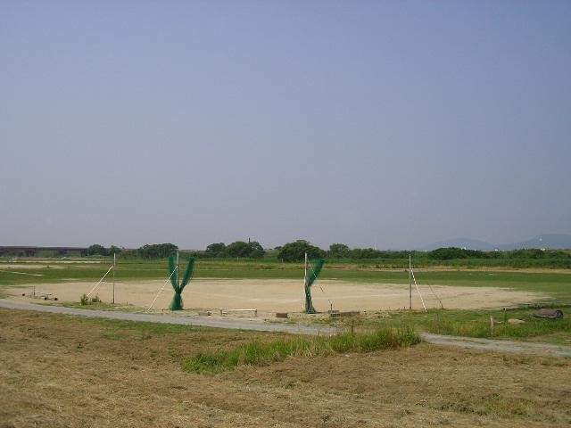 薄く霞んだ青空と遠方の山々を背景に、手前の小道から続く緑豊かな河川敷内に、防球ネットが設置された砂地の野球場や広場が複数整備されている様子を、少し高い位置から見渡すような構図で捉えた嘉瀬川河川敷北グラウンドの写真