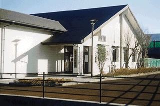 晴天の下、黒い三角屋根と白い外壁が特徴的な平屋建ての弓道場の建物を、手前の黒いフェンス越しに捉えた外観写真