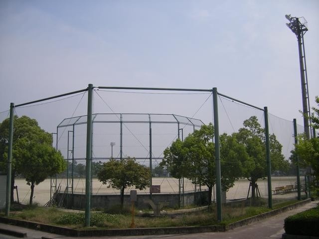 フェンスで囲われた野球場のバックネット裏付近からグラウンドを眺めた様子で、手前には緑豊かな木々が並び、右奥には高い照明塔が立っている佐賀市民運動広場の写真