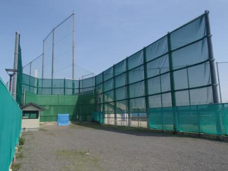 青空の下、砂利敷きの敷地を囲うように設置された、高い支柱と緑色の防球ネットが立ち、左側の小さな小屋とともに捉えた屋外弓道場の写真