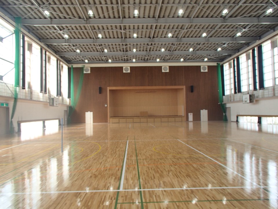 高い天井に鉄骨が剥き出しの構造を持つ広々とした体育館内部で、光沢のある木製フローリングにはバスケットボールやバドミントン用のラインが引かれ、正面の木目調の壁にはステージが設けられた体育センター内の写真