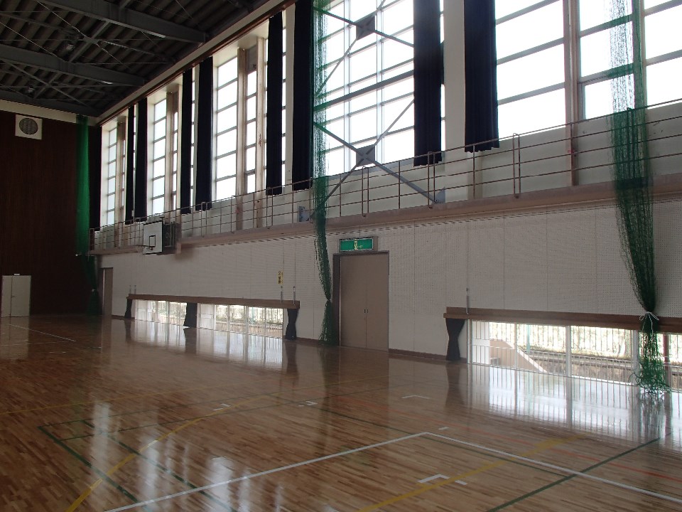 広々とした体育館の壁面に沿って、高い位置には黒いカーテンが掛かった大きな窓が並び、足元にも外光が差し込む小窓が配置され、磨き上げられた木製フローリングに窓からの光が美しく反射している体育センター内の写真