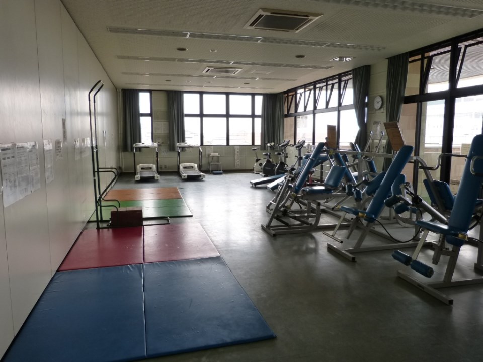 明るい窓から光が差し込むトレーニングルーム内に、数台のランニングマシンやエアロバイク、青いシートの筋力トレーニング器具が整然と並び、手前の床には色鮮やかなストレッチマットが敷かれた、清潔感のあるトレーニングハウスの写真