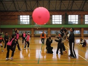 体育館で大きなピンク色のボールを囲み子どもと大人が走り回りながら協力して弾ませる様子を捉えた写真