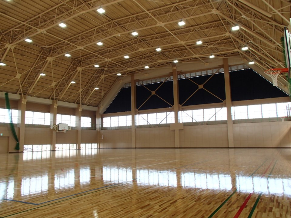 高い天井と鉄骨構造が広がる明るい室内で木目の床が広く輝くバスケットゴール付き体育館の写真