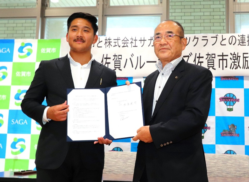 佐賀市と株式会社サガスポーツクラブとの連携を記念し、佐賀市長と代表者が並んで協定書を掲げながら、背後に佐賀市のロゴや「佐賀バルーナーズ佐賀市激励」の文字が記されたパネルを背に笑顔でポーズをとっている記念写真