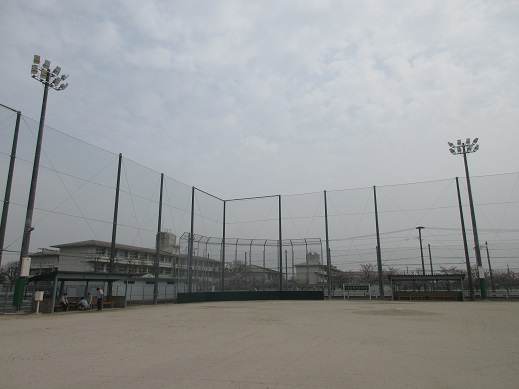 高い防球ネットに囲まれた砂地の野球場のグラウンドで、背後には3階建ての建物と照明塔が立ち並ぶ久保田グラウンドの写真