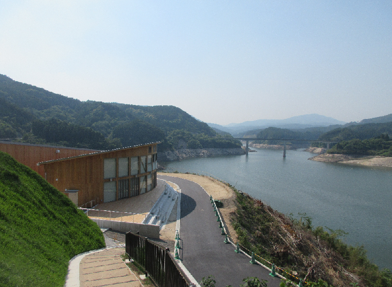 緩やかな曲線を描く木造の建物が、穏やかな湖と周囲の緑豊かな山々、そして遠くに架かる橋を見渡す高台に建っており、手前には湖畔へと続く舗装された道路と遊歩道の階段が整備されている水上競技場外観の写真