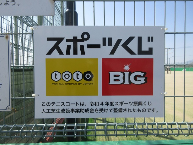 フェンスに掲示された看板に「スポーツくじ」と大きく記され、その下にtotoとBIGのロゴ、さらにこのテニスコートが令和4年度スポーツ振興くじ助成金を受けて整備された旨が書かれた、情報を伝達する写真