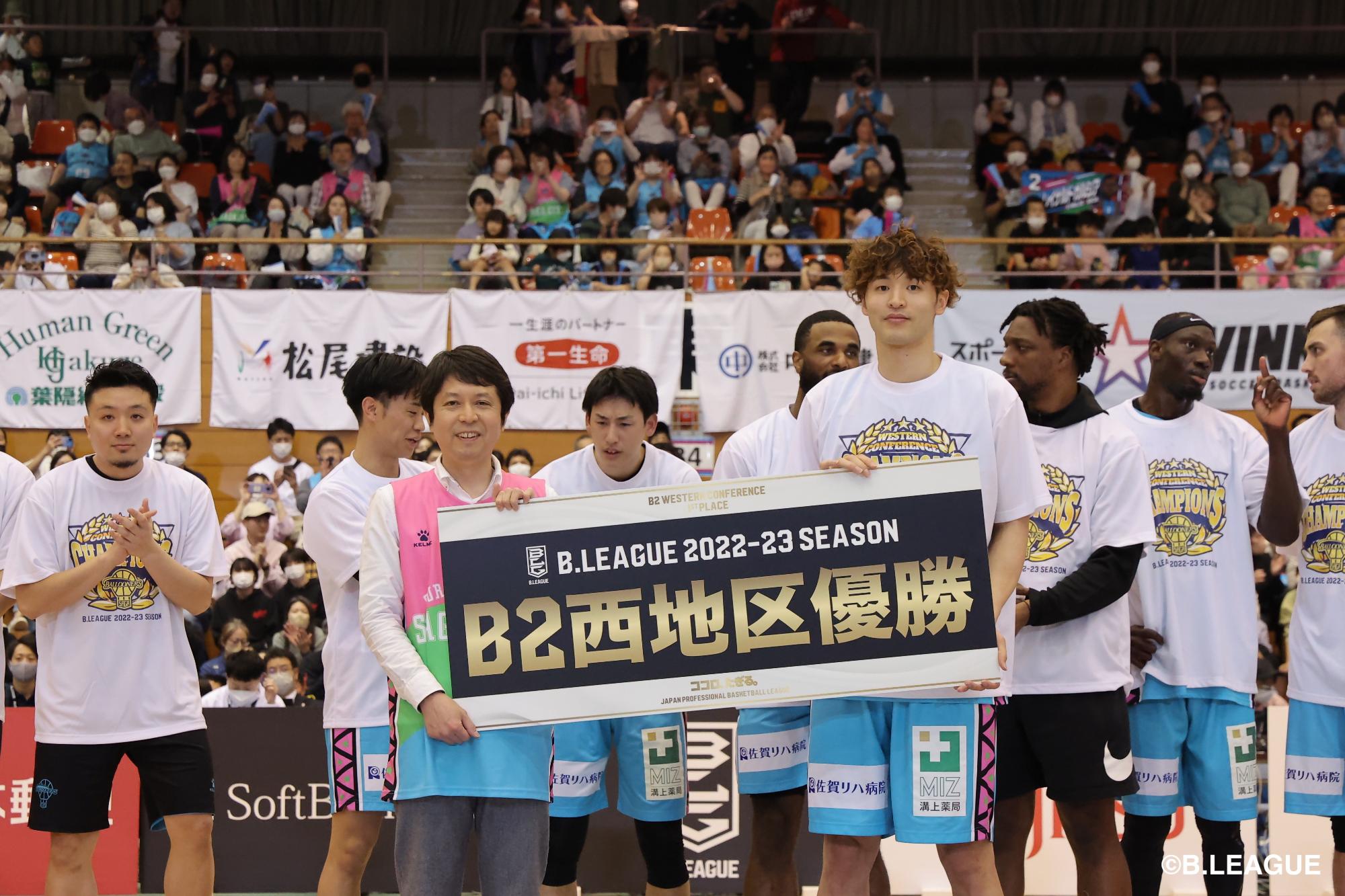 バスケットボールチームがコート上で大きなB2西地区優勝ボードを掲げ観客席を背に喜び合う様子の写真