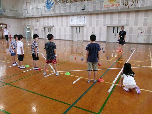 体育館で子どもたちが並んで指導者の前に立ち、色付きコーンやボールが置かれたエリアでボッチャをする様子を写した写真
