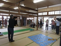 畳敷きの広間で複数の人々が輪になって立ち手を上げる動作を揃えて行う様子を写した写真