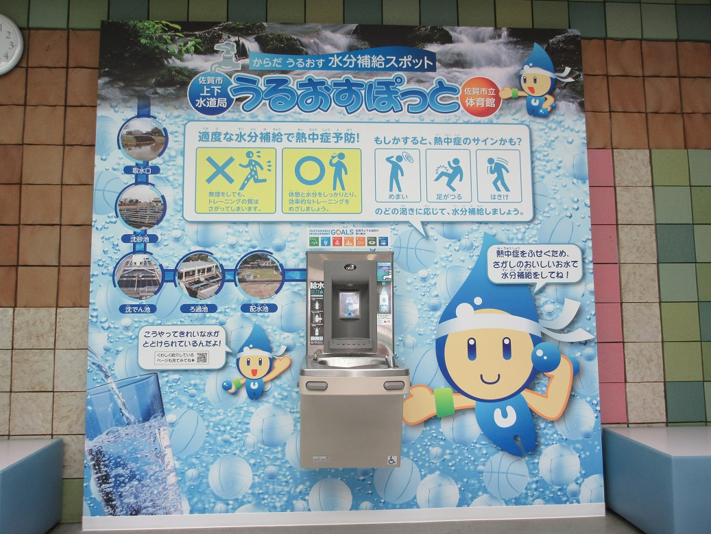 「うるおすぽっと」という愛称が書かれた青い掲示板の中央に冷水機が設置され、熱中症予防の重要性や水道水が届く仕組みをキャラクターと共に紹介している、体育館内の水分補給スポットを正面から捉えた写真